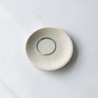 A minimalist ceramic dish with a textured cream-beige glaze, featuring a thin blue teal wire bracelet inside its shallow center, set against a clean white background.