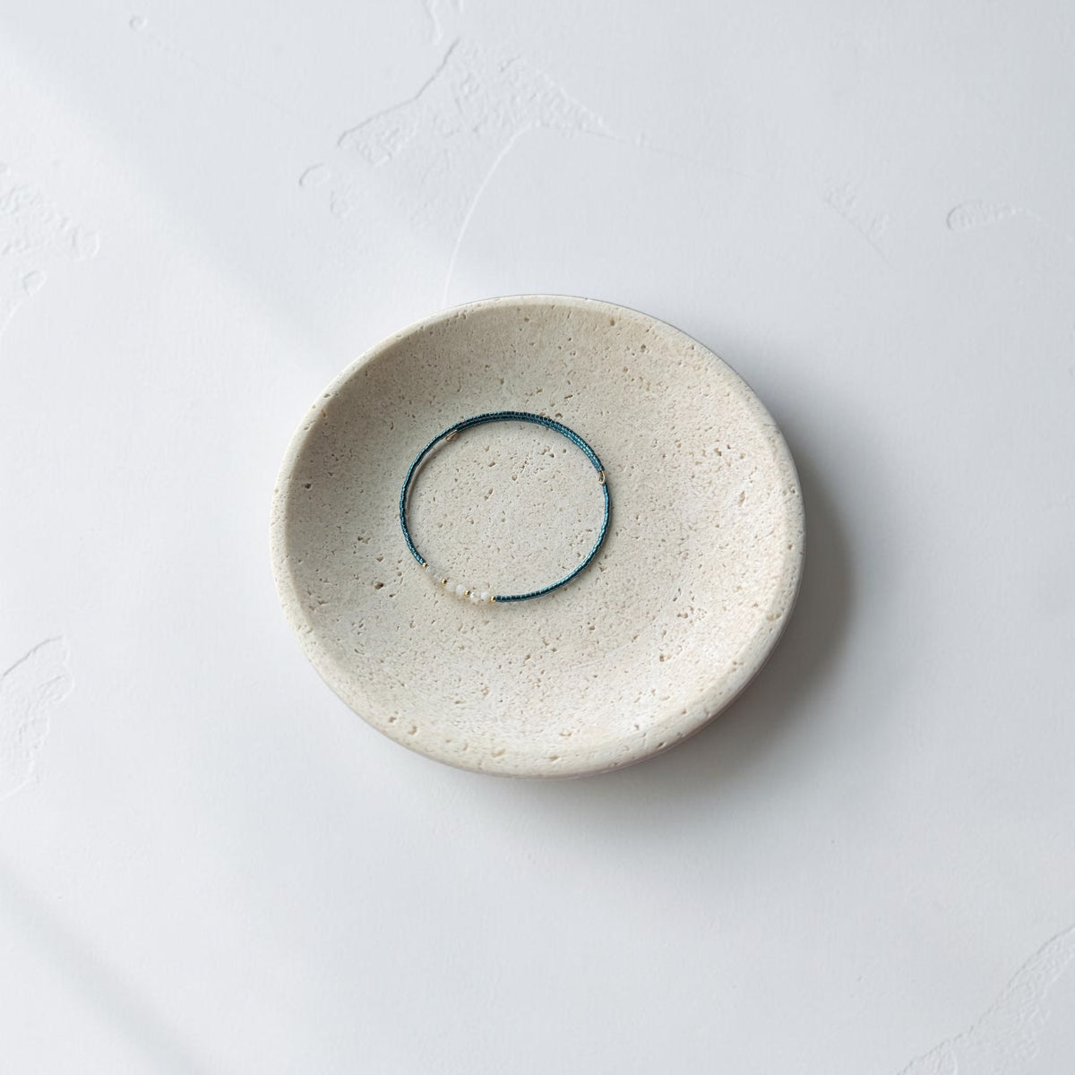 A minimalist ceramic dish with a textured cream-beige glaze, featuring a thin blue teal wire bracelet inside its shallow center, set against a clean white background.