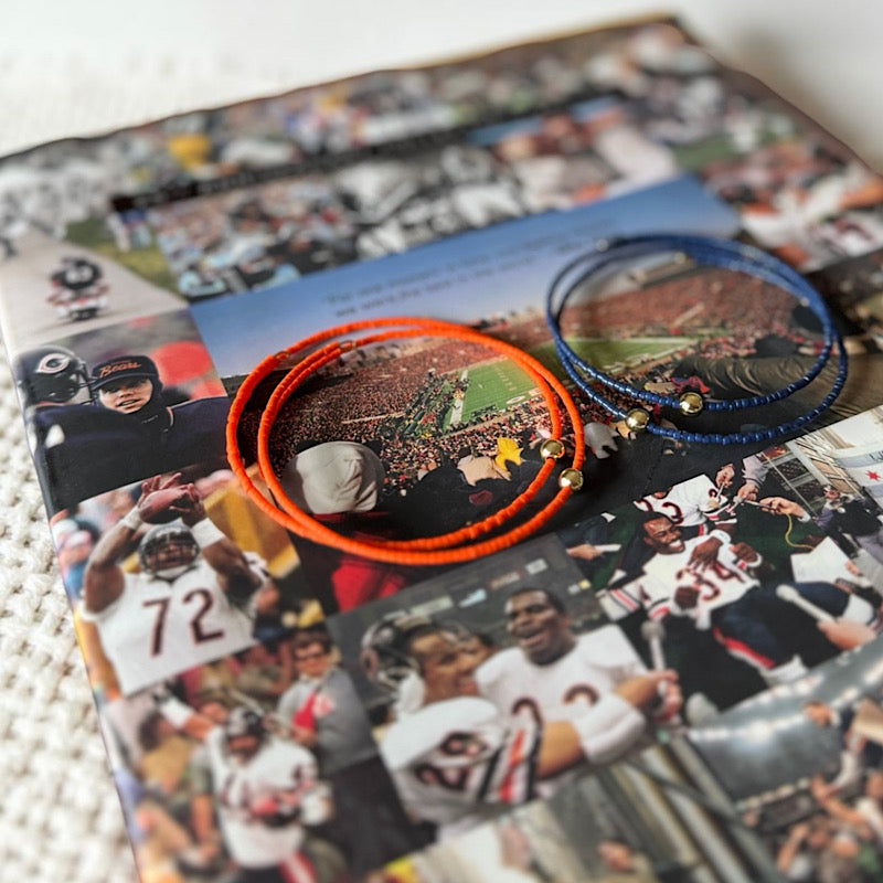 Two handmade wire bracelets, one bright orange and one blue, displayed on top of a custom collage print featuring football players and stadium scenes.