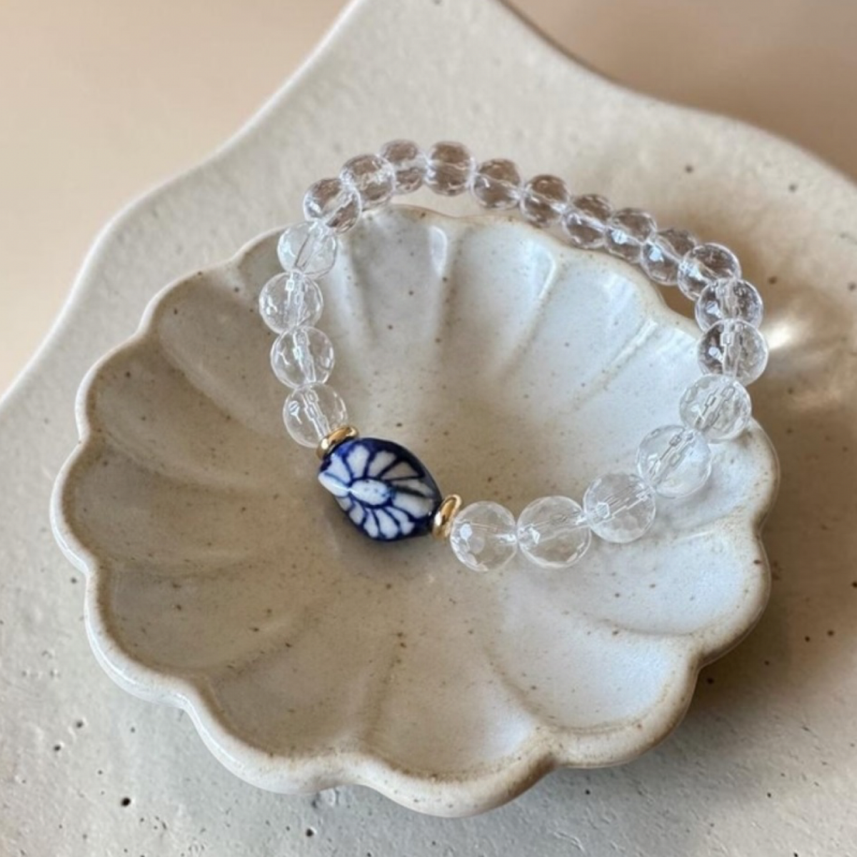 A beaded stretch bracelet with clear faceted beads and a central blue-and-white floral bead with gold-toned metal, displayed on a scalloped cream ceramic dish.