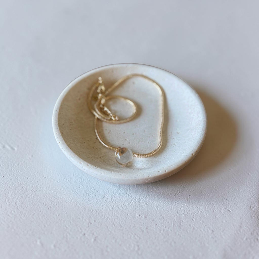 A delicate gold chain necklace with a small clear stone pendant, displayed inside a small round beige ceramic dish on a textured light gray surface.