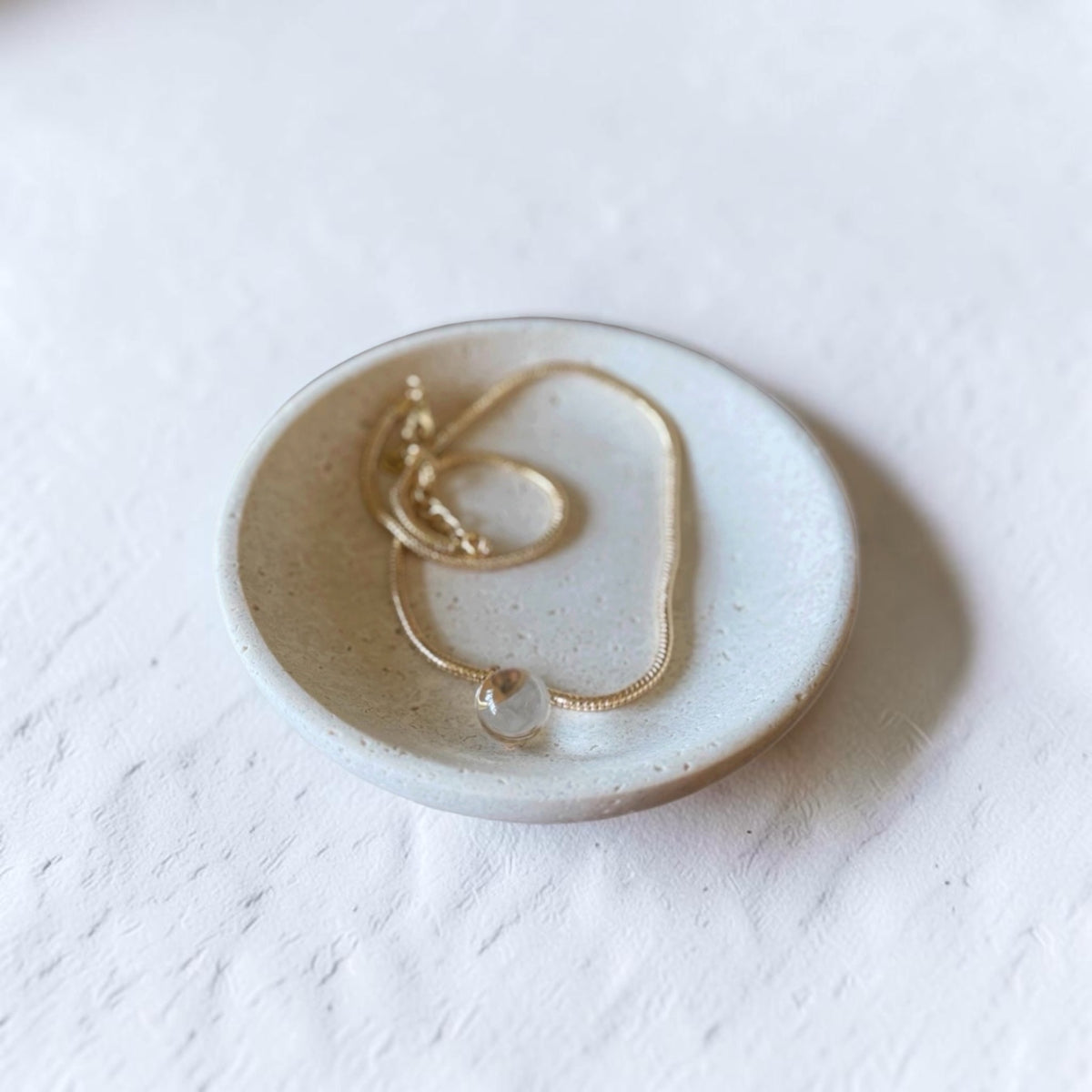 A delicate gold chain necklace with a small clear bead pendant, displayed inside a small round ceramic dish on a textured white surface.