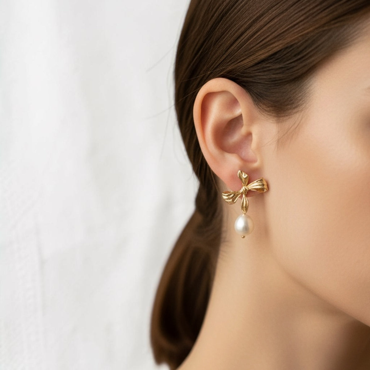 A close-up of a woman's ear wearing a gold bow-shaped stud earring with a dangling white pearl, against a soft white background.
