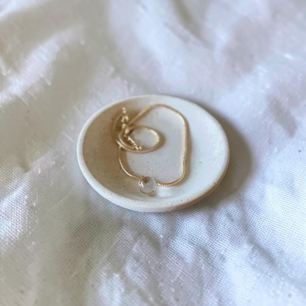 A delicate gold chain necklace with a small clear bead pendant rests on a small round ceramic dish, set against a soft white textured fabric background.