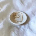 A delicate gold chain necklace with a small clear bead pendant rests on a small round ceramic dish, set against a soft white textured fabric background.