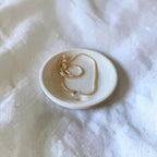 A delicate gold chain necklace with a small clear bead pendant rests on a small round ceramic dish, set against a soft white textured fabric background.