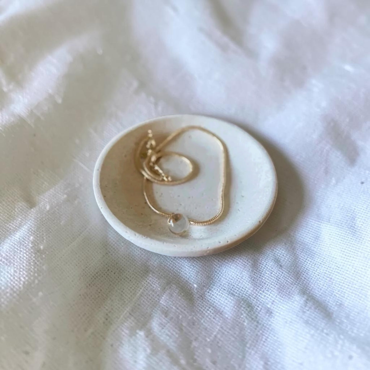 A delicate gold chain necklace with a small clear bead pendant rests on a small round ceramic dish, set against a soft white textured fabric background.