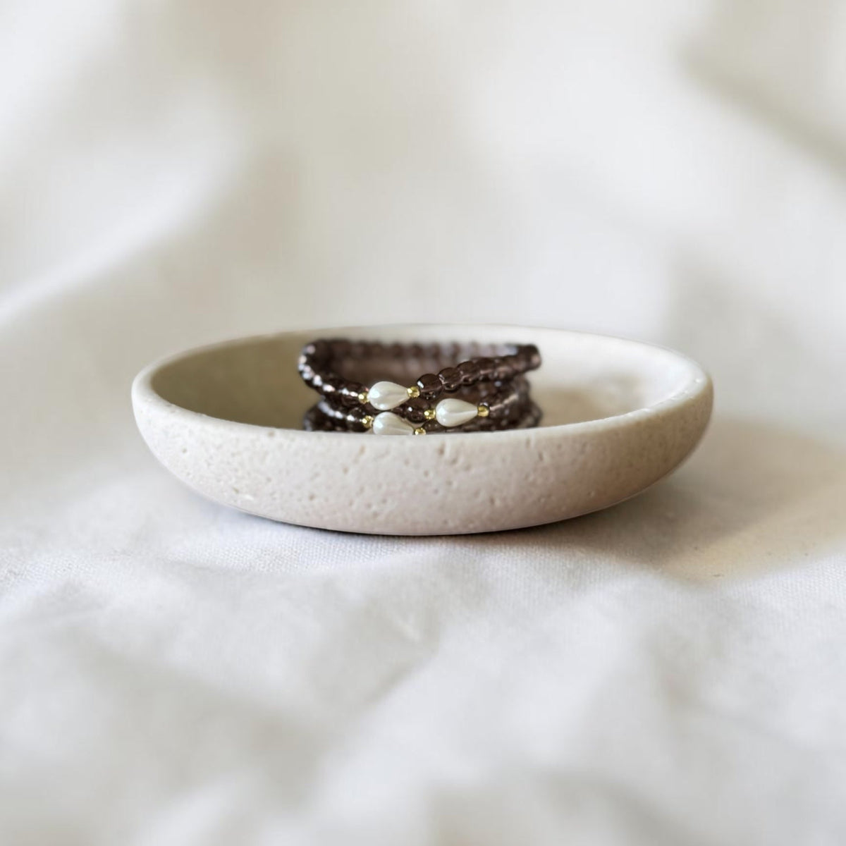 A close-up of a small round ceramic dish holding three dark brown beaded bracelets with white pearl-like accents, set against a softly blurred white fabric background.
