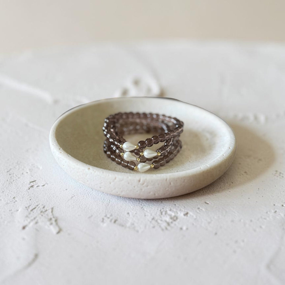 A stack of three beaded wrap bracelets in a dark brown smoky color with small white pearl-like beads, displayed on a small round ceramic dish.