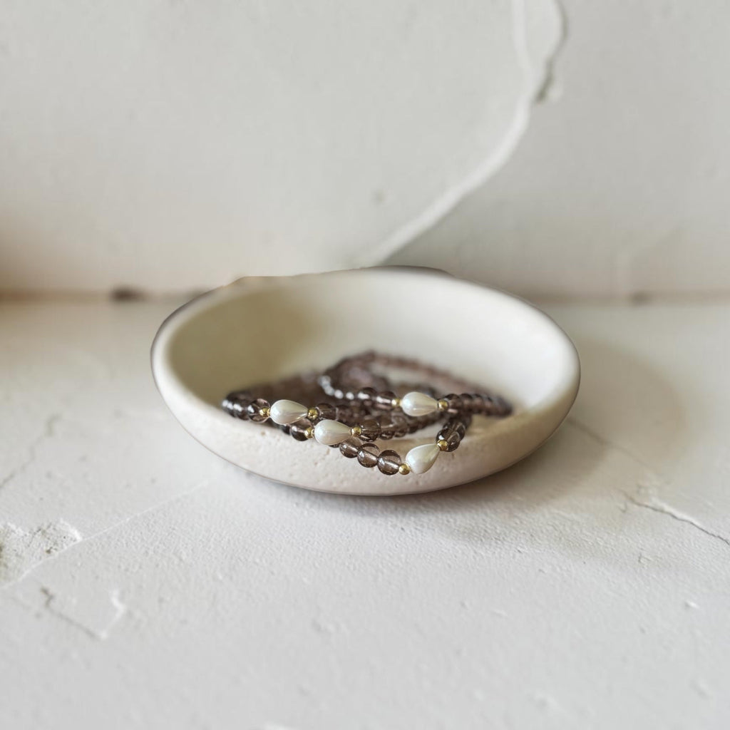 A small round ceramic dish holding several strands of beaded jewelry, photographed against a textured off-white wall and surface.