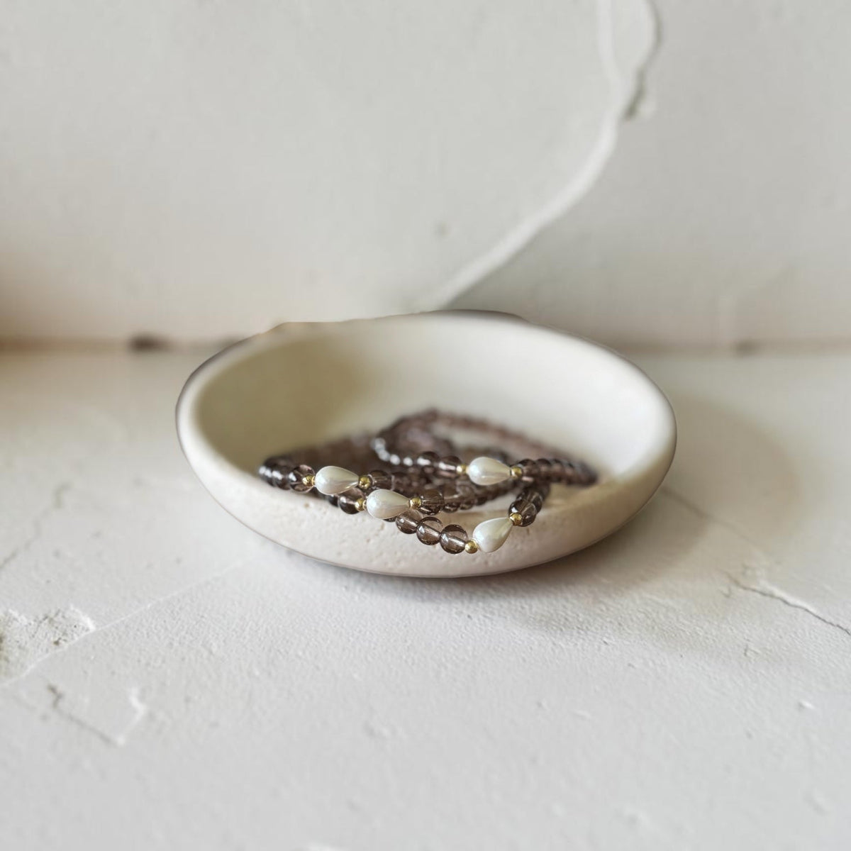 A small round ceramic dish holding several strands of beaded jewelry, photographed against a textured off-white wall and surface.