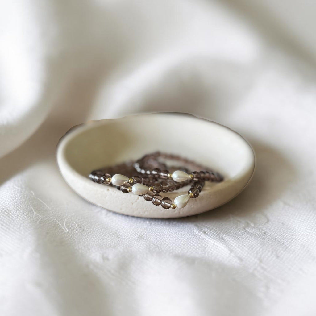 A small round ceramic dish holding a beaded necklace with white pearl-like beads and dark brown beads, set against a soft white fabric background.
