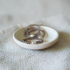 A small round off-white ceramic dish holding several beaded rings, set on a textured cream fabric against a soft blurred light background.
