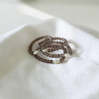 A stack of beaded bracelets with dark brown translucent beads and small white pearl accents, resting on a soft white fabric surface.