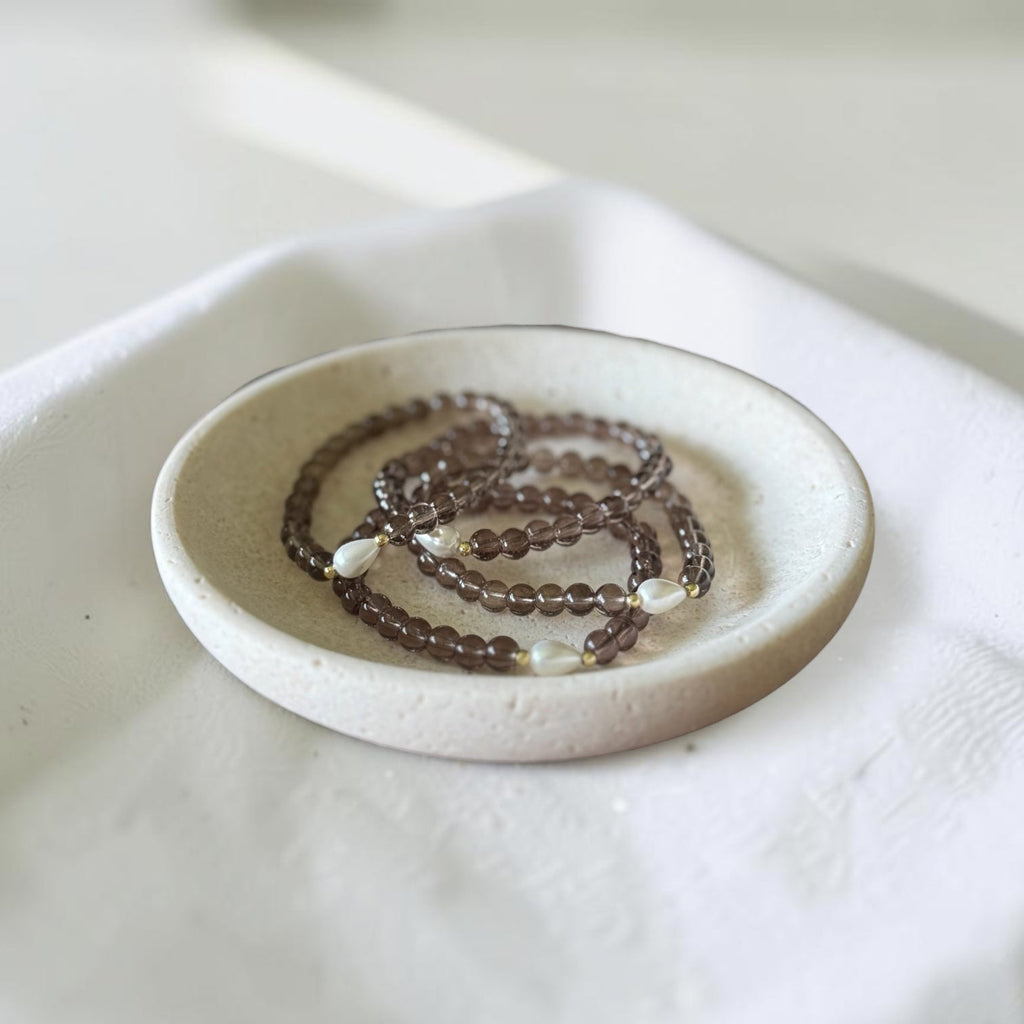 A beaded necklace with dark brown translucent beads and small white pearl-like accents, displayed in a shallow round ceramic dish on a white fabric surface.