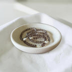 A beaded necklace with dark brown translucent beads and small white pearl-like accents, displayed in a shallow round ceramic dish on a white fabric surface.