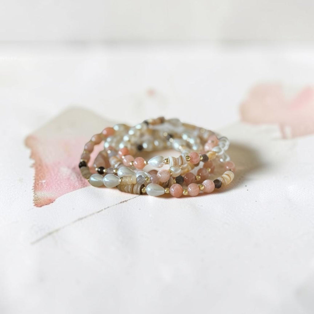 A stack of multi-strand beaded rings in soft pastel colors, including pink, beige, gray, and white, arranged on a white surface with a soft pink watercolor accent.