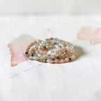 A stack of multi-strand beaded rings in soft pastel colors, including pink, beige, gray, and white, arranged on a white surface with a soft pink watercolor accent.
