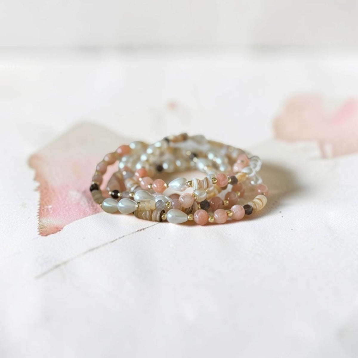 A stack of multi-strand beaded rings in soft pastel colors, including pink, beige, gray, and white, arranged on a white surface with a soft pink watercolor accent.