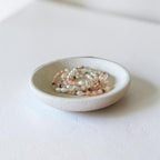 A small round ceramic dish holding several strands of beaded bracelets in soft pastel colors, photographed on a clean white surface with a softly blurred neutral background.