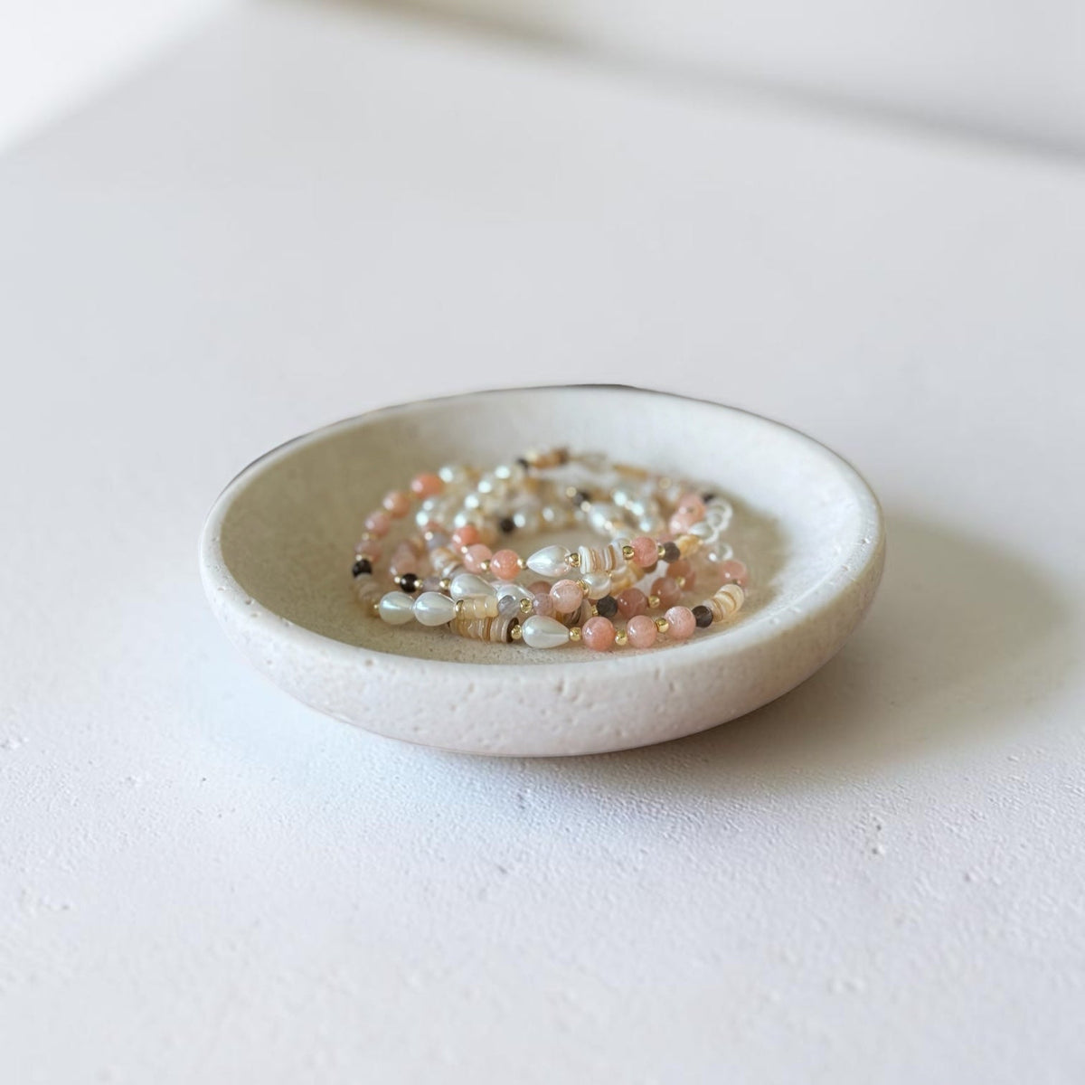A small round ceramic dish holding several strands of beaded bracelets in soft pastel colors, photographed on a clean white surface with a softly blurred neutral background.