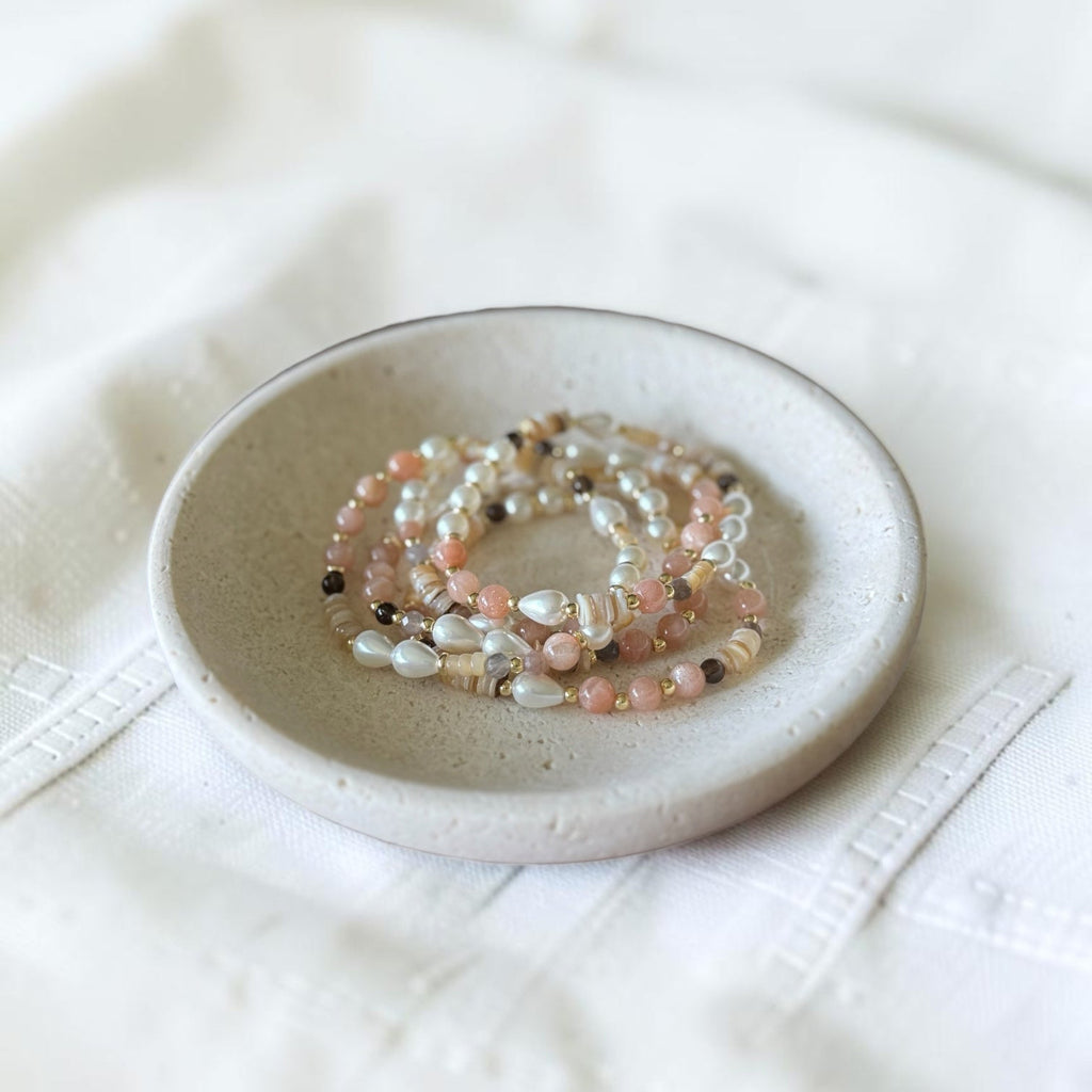 A stack of multi-strand beaded bracelets in soft pink, cream, and white tones, displayed on a small speckled ceramic dish over a textured white fabric background.