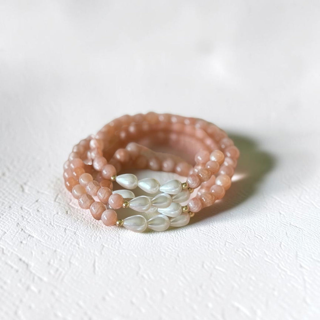 A multi-strand beaded bracelets set in a dusty pink tone with a central cluster of white teardrop-shaped beads, photographed on a textured white surface.