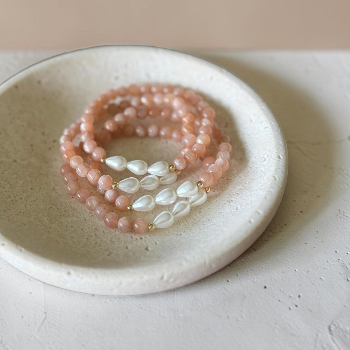 Three beaded stretch bracelets displayed on a small round ceramic dish. Two bracelets are made of peach-pink beads, and one is made of white baroque pearls with small gold spacer beads.