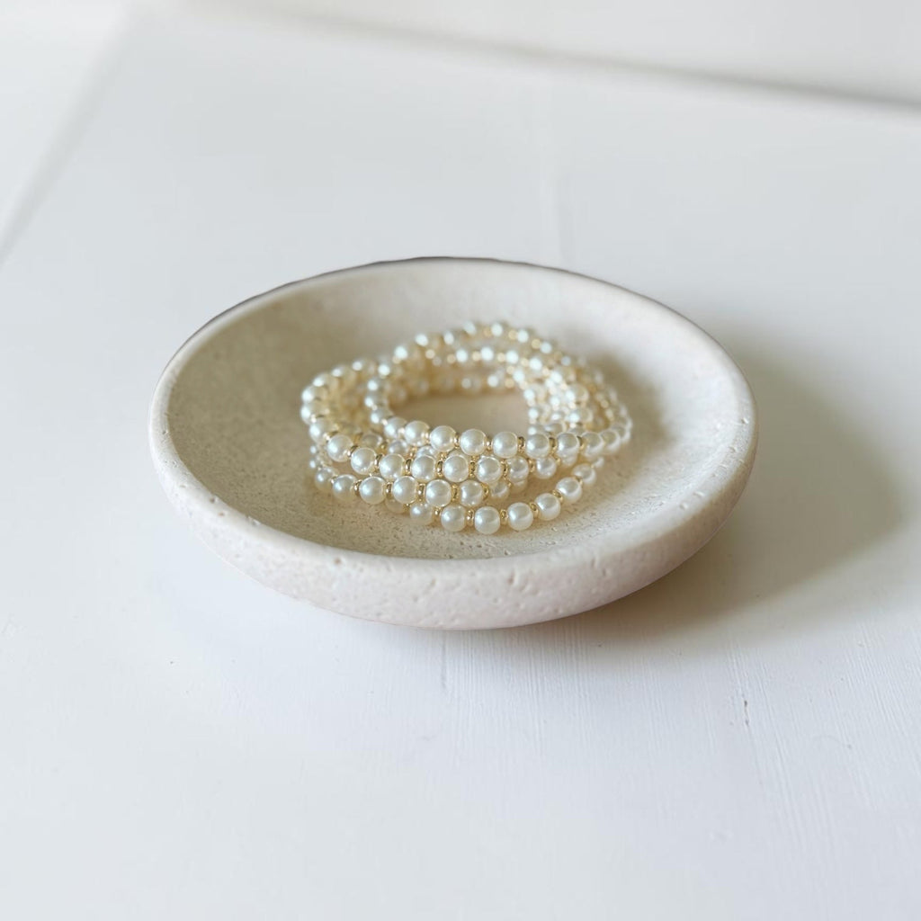 A stack of pearl-strand bracelets displayed in a shallow round ceramic dish on a clean white surface.