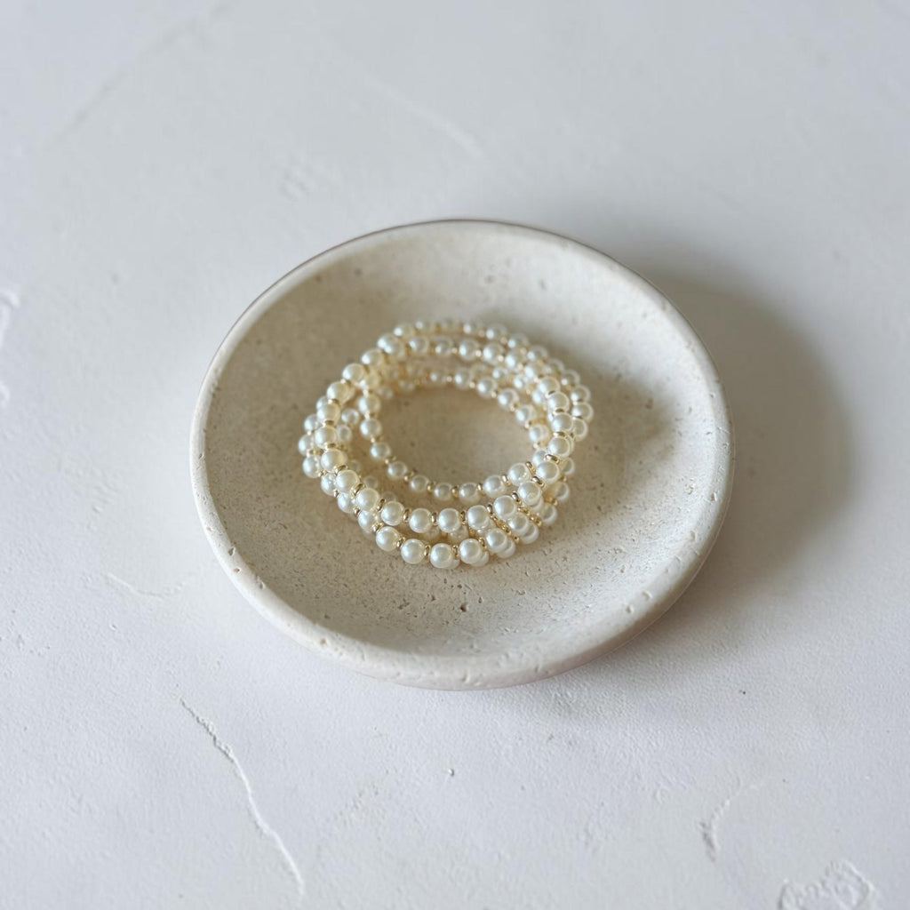 Three strands of white faux pearl beaded bracelets stacked on a small round ceramic dish, set against a textured light gray background.