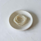 Three strands of white faux pearl beaded bracelets stacked on a small round ceramic dish, set against a textured light gray background.
