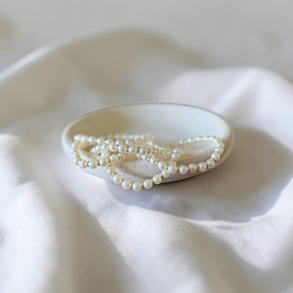 A strand of small white pearl-like beads draped over a small oval dish on soft white fabric.