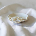 A strand of small white pearl-like beads draped over a small oval dish on soft white fabric.
