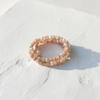 A delicate beaded ring with small iridescent pearls and pale pink beads, resting on a textured white surface with a soft shadow.