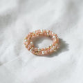 A delicate double-strand beaded ring featuring light pink beads, white pearl-like beads, and small gold spacer beads, resting on a textured white fabric background.