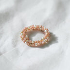 A delicate double-strand beaded ring featuring light pink beads, white pearl-like beads, and small gold spacer beads, resting on a textured white fabric background.