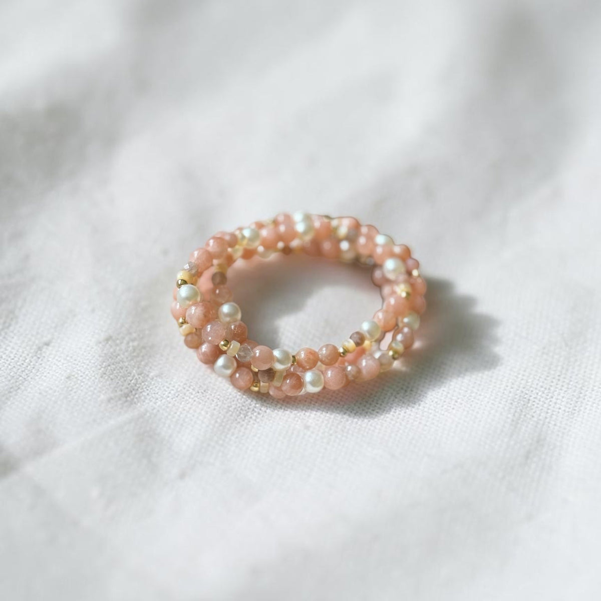 A delicate double-strand beaded ring featuring light pink beads, white pearl-like beads, and small gold spacer beads, resting on a textured white fabric background.