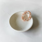 A peach and white beaded stretch bracelet displayed on a small speckled ceramic dish against a light gray background.
