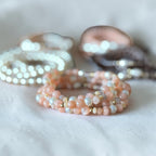 A close-up of a beaded stretch bracelet in soft peach-pink and iridescent white beads, with other blurred jewelry in the background on a white fabric surface.