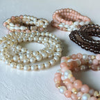 A group of beaded stretch bracelets arranged on a textured white surface. The bracelets feature white faux pearls with gold-toned spacers, peach-pink beads with gold accents, and dark brown beads.
