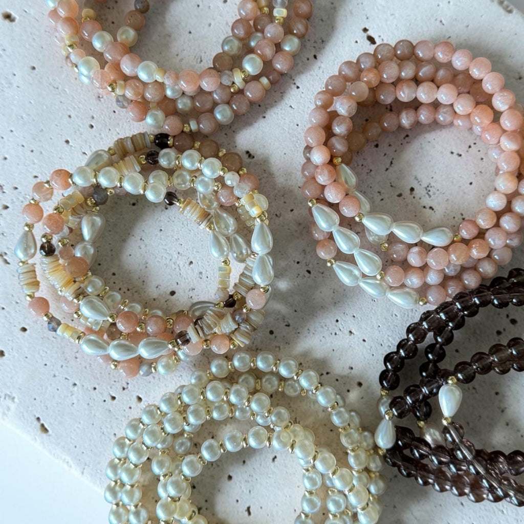 Six beaded stretch bracelets arranged on a speckled white stone slab. The bracelets feature pastel pink, cream, white, and dark brown beads, with some showing iridescent pearl-like accents.