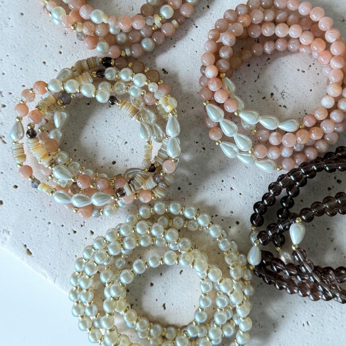 Six beaded stretch bracelets arranged on a speckled white stone slab. The bracelets feature pastel pink, cream, white, and dark brown beads, with some showing iridescent pearl-like accents.