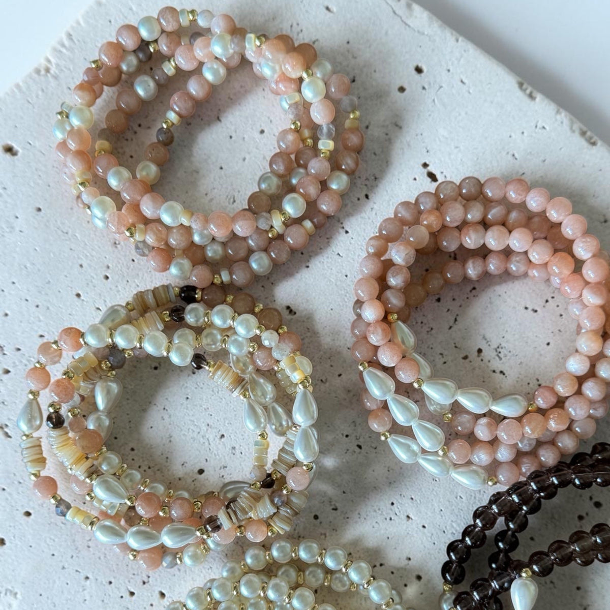 Six beaded stretch bracelets arranged on a speckled white stone slab. The bracelets feature pastel pink, cream, white, and dark brown beads, with some showing iridescent pearl-like accents.