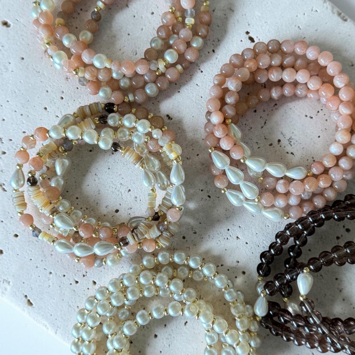 Six beaded stretch bracelets arranged on a speckled white stone slab. The bracelets feature pastel pink, cream, white, and dark brown beads, with some showing iridescent pearl-like accents.