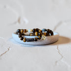 Four beaded stretch bracelets with brown tiger's eye and dark beads, displayed on a small white ceramic dish against a textured light beige background.