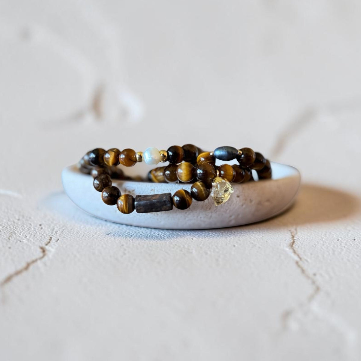 Four beaded stretch bracelets with brown tiger's eye and dark beads, displayed on a small white ceramic dish against a textured light beige background.