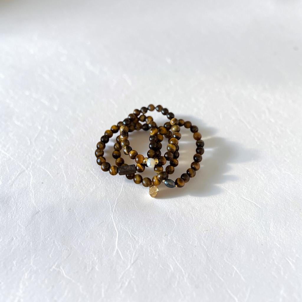 A set of beaded stretch bracelets in shades of brown, black, and gold, arranged in a small coil on a textured white surface.