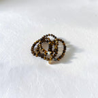 A set of beaded stretch bracelets in shades of brown, black, and gold, arranged in a small coil on a textured white surface.