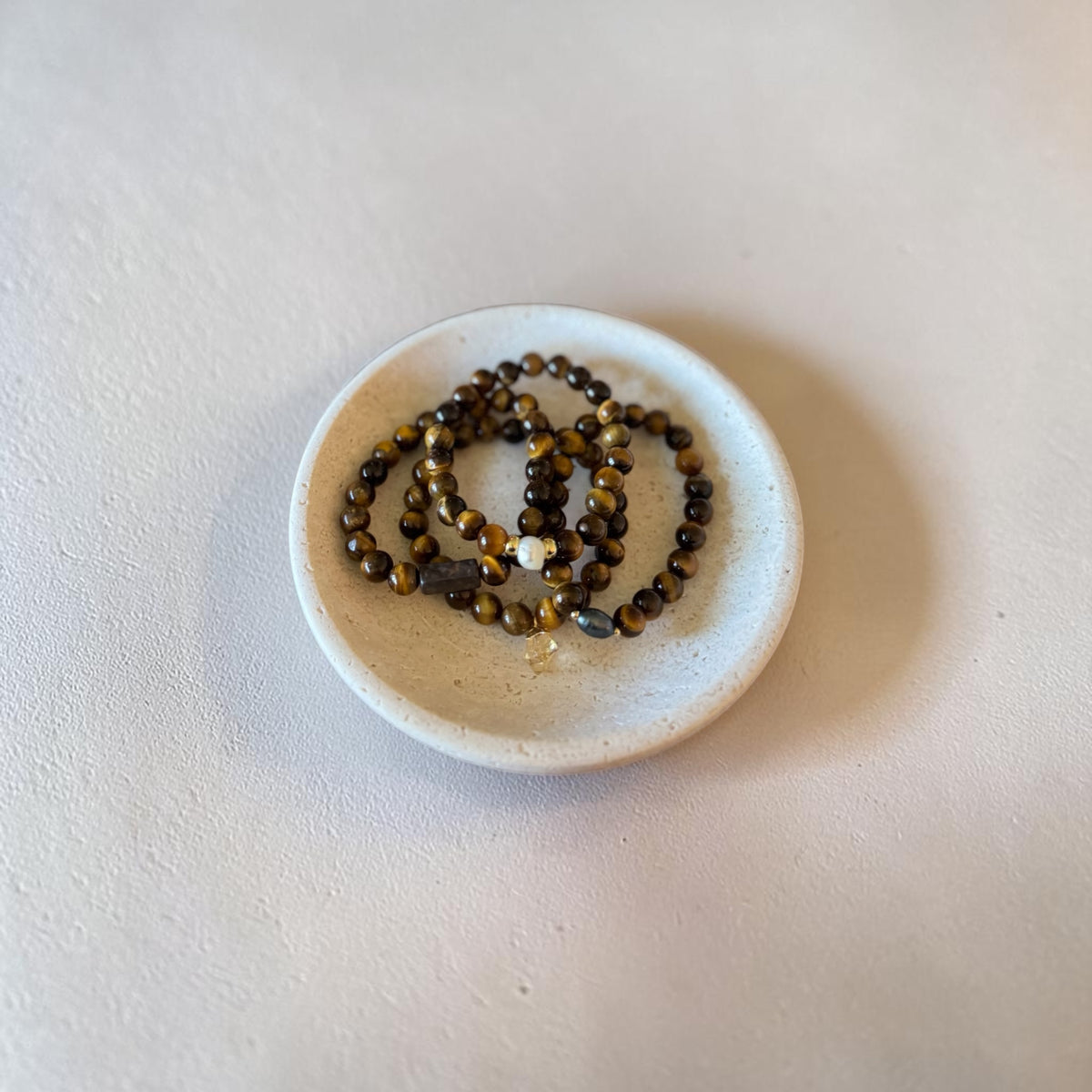 Four beaded bracelets with brown and gold tiger's eye beads and a small white bead, displayed in a small white ceramic dish on a textured beige surface.