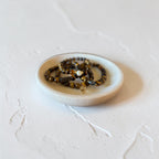 A small round ceramic dish holding two beaded bracelets with brown, black, and gold-toned beads, photographed on a textured off-white surface.